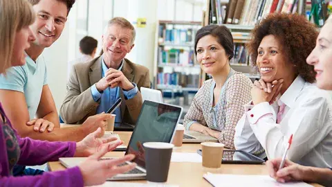 A group of adult learners smiling and practicing vocabulary and writing together in a library classroom.