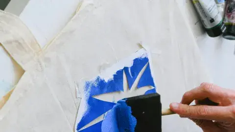 Hand brushing a sponge brush with blue paint over a pointed stencil shape on a natural colored tote bag on a white table.