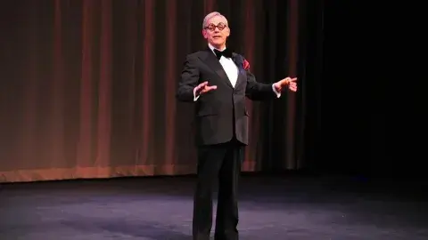 A performer holds the signature cigar, and wears the tuxedo and glasses of famed comedian George Burns