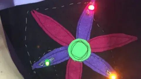 Alternating pieces of magenta and purple petal-shaped felt with a neon green circle piece sewed on a black square of felt with green, pink, and orange LED lights.