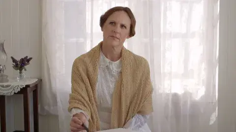 Molly Shannon dressed as Emily Dickinson sits in a sunlight room holding a pen