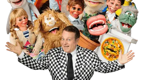 Smiling puppeter is surrounded by various puppets. 