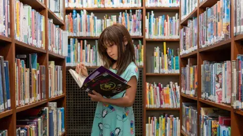 Head & shoulder view of brunette, white girl, reading book her in arms, surrounded by book-filled shelves
