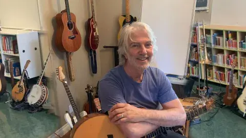 Smiling white-haired white male in blue shirt holding a guitar