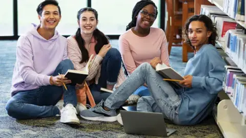 Group of four high school aged students sitting together with notebooks and a laptop on the floor of the library and studying.