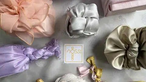 a table decorated with bojagi gift wrappings in various shades of pink, purple, grey, and yellow with a card that says Nossi