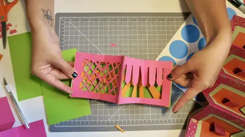 hands displaying pop up book art project, scissors and paper on table