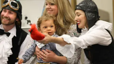 Two LA Opera performers in costume holding a bird prop in front of a young mother with her baby toddler.