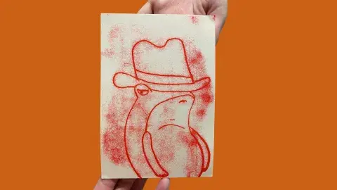 hands holding a mono print of a red frog wearing a cowboy hat