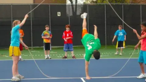 People holding a jump rope on a tennis court, man in center doing a handstand