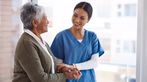 older adult dressed in a brown sweater walking with nurse