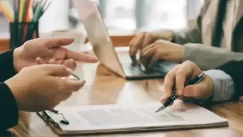 The hands of 3 people at a table signing a document and typing on a laptop