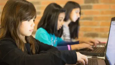 three female students sitting in a row with ling brown hair working on computers. there is a red brick background and a bright light on their faces from their devices. 