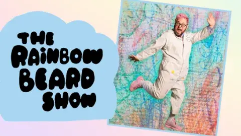 A photo of Ricky Rainbow Beard, with pink hair, a jumpsuit, and pink shoes, dancing in front of a colorful background that looks like crayon scribbles