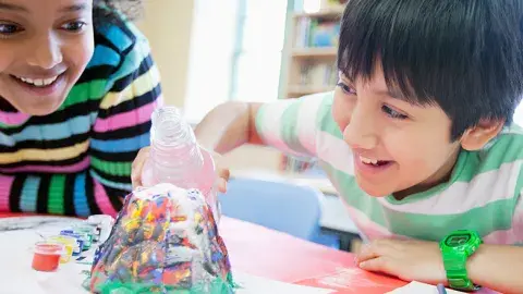 Two smiling children bent over a volcano science experiment pouring in the solution and waiting for it to bubble over.