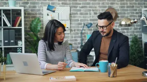 A man and woman sitting at a desk with work materials, looking at a document together