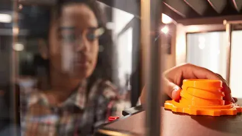 A hand reaching into a 3D printer to touch a 3D printed object on the print bed. 