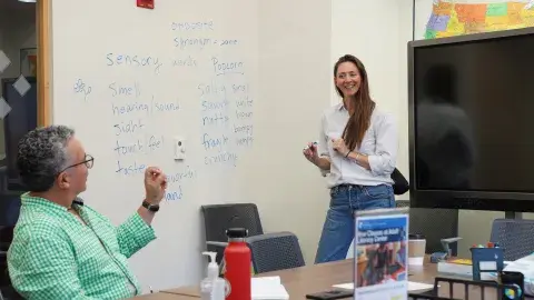 teacher in white blouse and blue jeans long brown hair teaching a learner with grey hair and a light green dress shirt. 