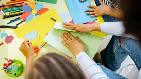 An assortment of craft supplies, paints, markers and hands with scissors cutting paper.