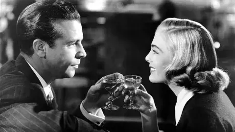 The protagonists, a man and a woman, are looking at each other smiling and holding glasses with alcoholic drinks