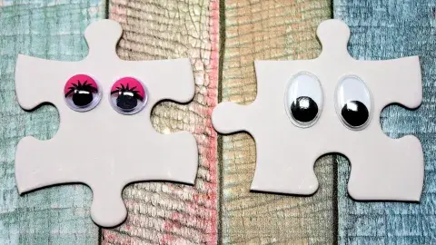 Two white jigsaw puzzle pieces side by side decorated with wiggle eyes against a backdrop of different colored stained wood.