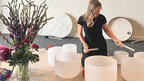 A woman sitting in the middle of six variously sized glass bowls, looking down with the playing sticks.