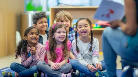 5 small smiling children sitting on a rug facing an adult holding a book. 