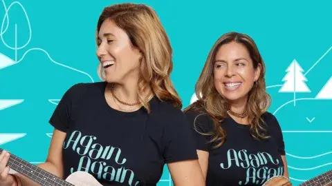 Two smiling women with guitars, wearing t-shirts that say Again Again