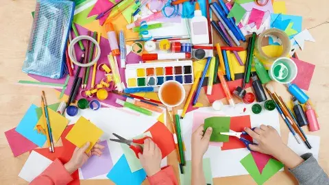 Children's hands holding scissors and cutting paper with a table full of arts and crafts supplies