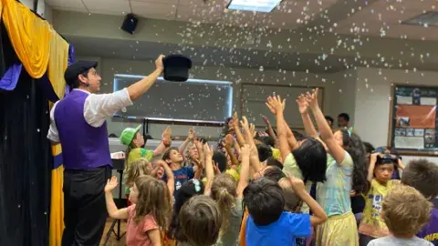 A man in a purple vest, standing in front of a yellow curtain, holding up a black top hat. There are bubbles coming out of the hat, and a crowd of young children are reaching up towards the bubbles.