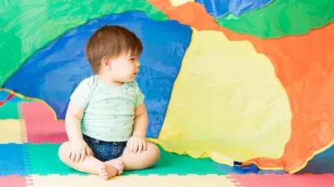 Baby sits on a colorful floor in front of an equally colorful parachute.