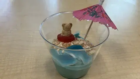 A cup of blue pudding on a table decorated with cookie crumbs, candy and a paper umbrella