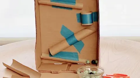 Cardboard rolls taped to a larger cardboard rectangle with a bowl of marbles in front 