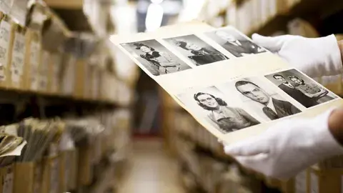 In the foreground are white gloved hands holding a white sheet with black-and-white photographs of people. In the background are rows of boxes on shelves.