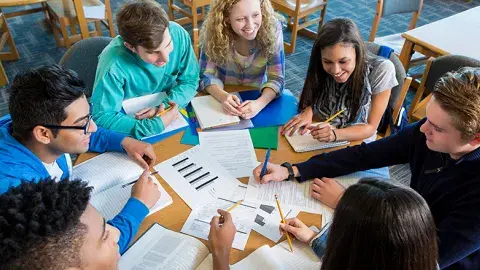 A group of seven teenage girls and boys seated around a round table with documents, notebooks and opened textbooks scattered on the surface.