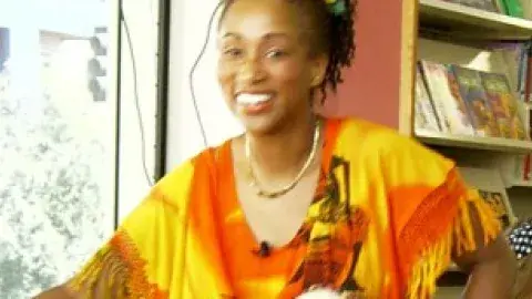 Ina in yellow and orange robes holding a jimbe drum.