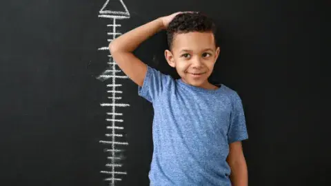 a young boy stands in front of a line of chalk height measurements on a black wall