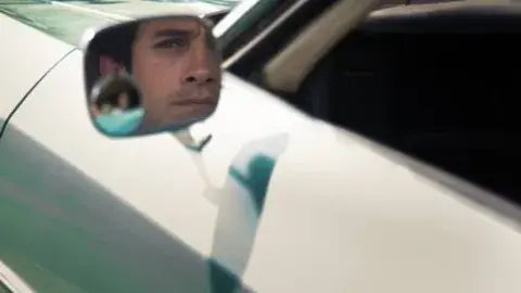 A drivers side-view mirror captures the reflection of a character played by Gael García Bernal.