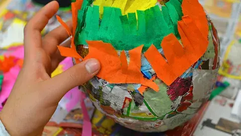 A child’s hand applies cut tissue paper to a paper mache form.