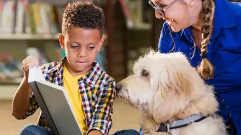 Young boy holding up a book for a shaggy dog to look at.