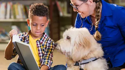 a young boy shows a book to an attentive dog and its adult handler