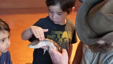 a man in a hat holds a lizard while two boys look