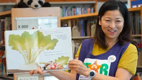 A woman woman wearing a STAR volunteer vest holds up a picture book in a library