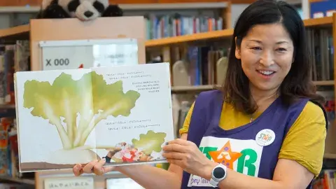 A woman woman wearing a STAR volunteer vest holds up a picture book in a library