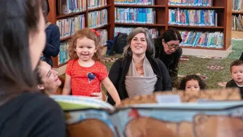 Several children and adults watch a woman with long dark hair holding a picture book.