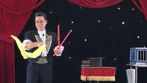 A magician holding wands and a scarf on stage next to a table with a top hat and a cage