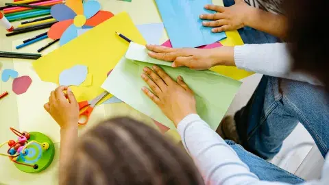 An assortment of craft supplies, paints, markers and hands with scissors cutting paper.