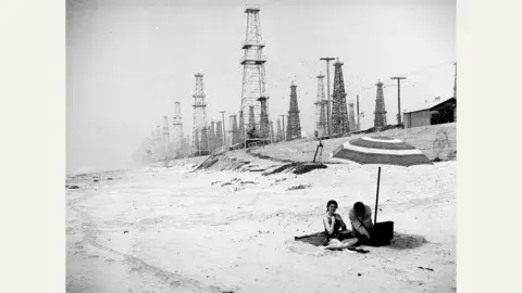 A couple spread out a picnic in the shadows of Huntington Beach oil derricks, circa 1937
