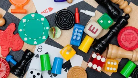Assorted board game pieces, poker chips, dice, chess pieces, playing cards, and letter blocks scattered on a checkered surface.