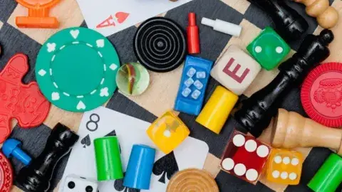 Colorful board-game pieces, cards, dice, chips, pawns, and wooden tokens on a tabletop scene on a checkered background.